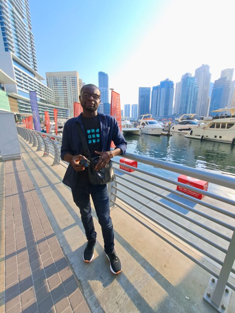 Kojo standing along the Dubai Marina waterfront with yachts and skyscrapers in the background.