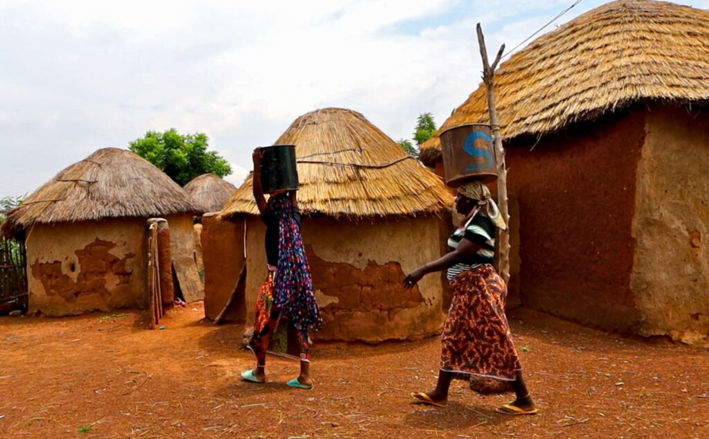 Two Ghanaian women carry buckets of water on their heads, walking past traditional round mud huts with thatched roofs in a rural village.