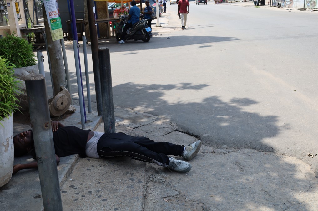 A man lying on the sidewalk in Osu, Accra, appearing exhausted or unwell