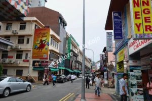 A lively street view of Little India, Singapore, with colorful shops, people walking, and cars passing through.
