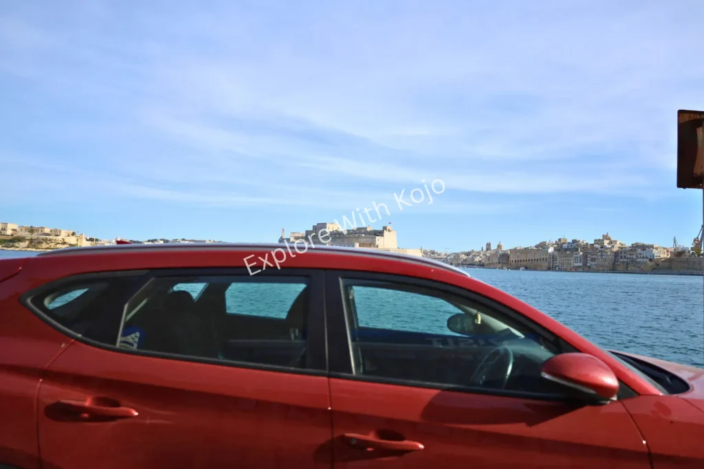 Red car parked by the harbor in Malta with waterfront in view.