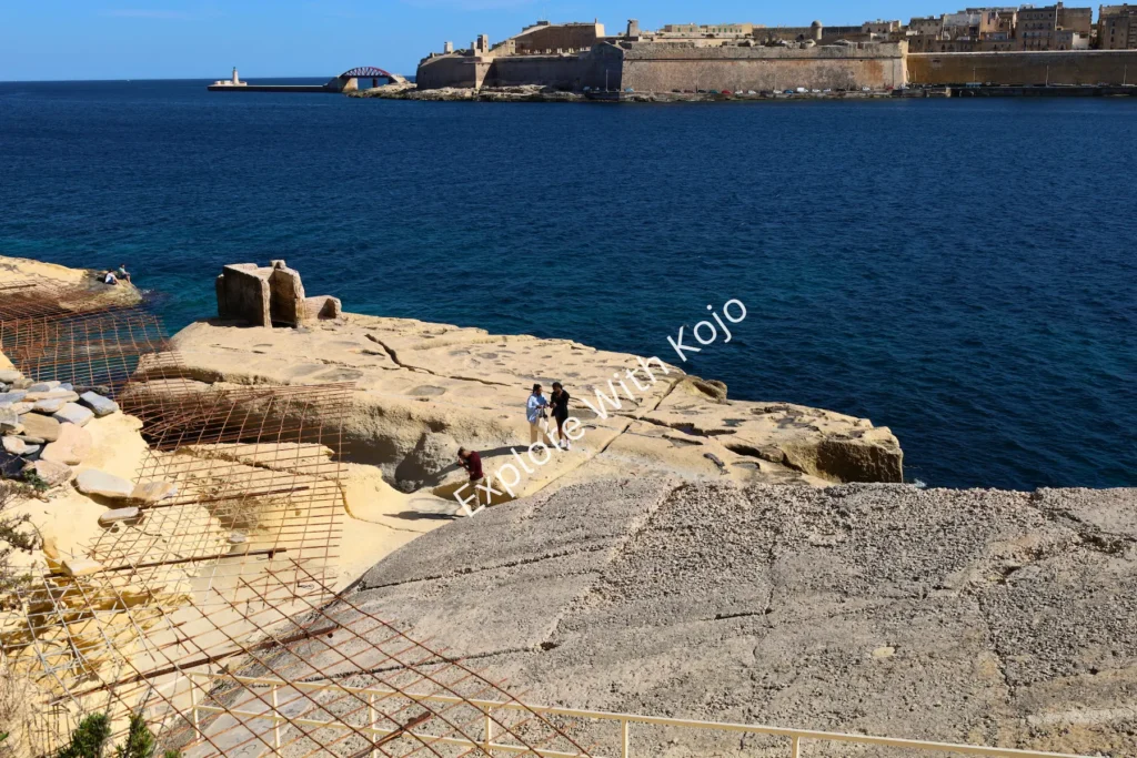 Rocky jetty in Malta extending into the sea with fortifications in&nbsp;the&nbsp;distance.