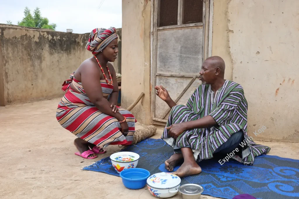 African man speaks with his second wife as she kneels to clear his finished meal, showing deep respect and humility in a traditional courtyard setting.