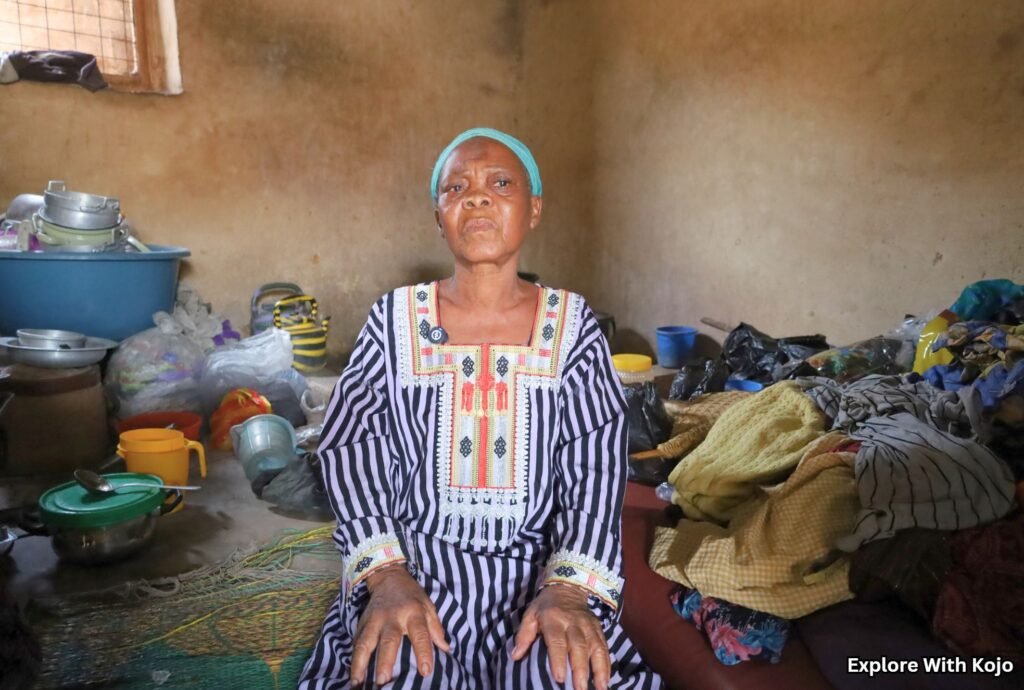 Elderly Ghanaian woman sitting indoors in a modest home, surrounded by household items and clothing.