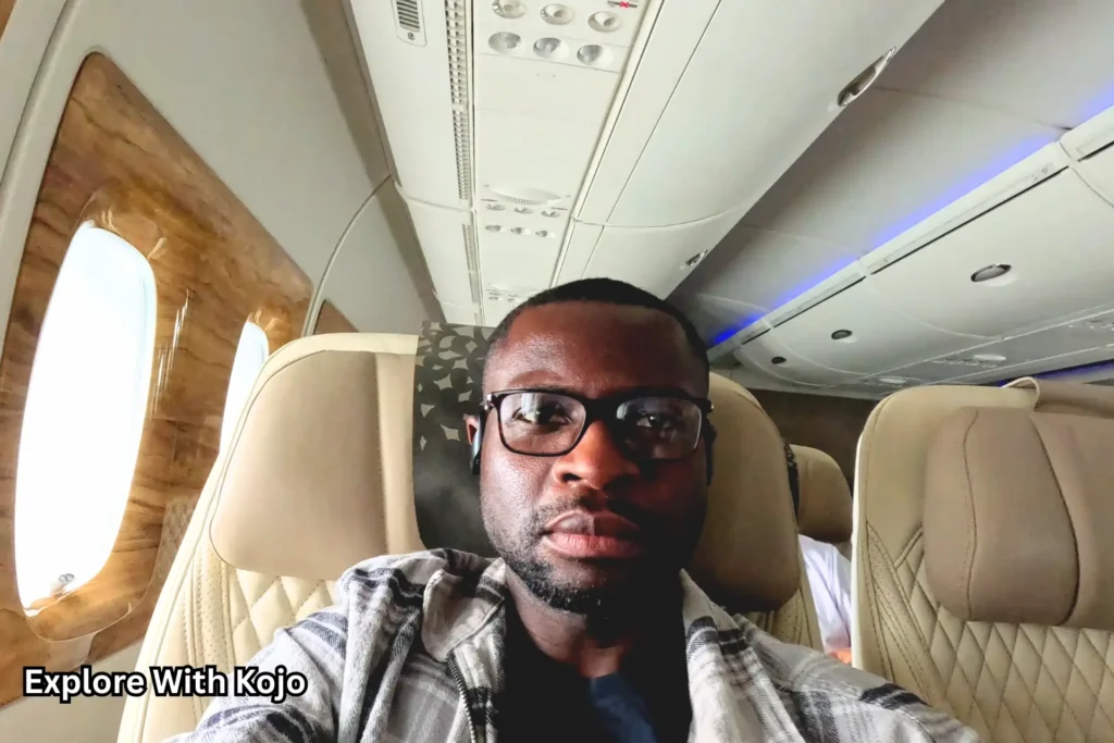 Kojo seated in Emirates Premium Economy, close-up selfie showing cream leather seat, cabin ceiling panels and inflight screen in the background.