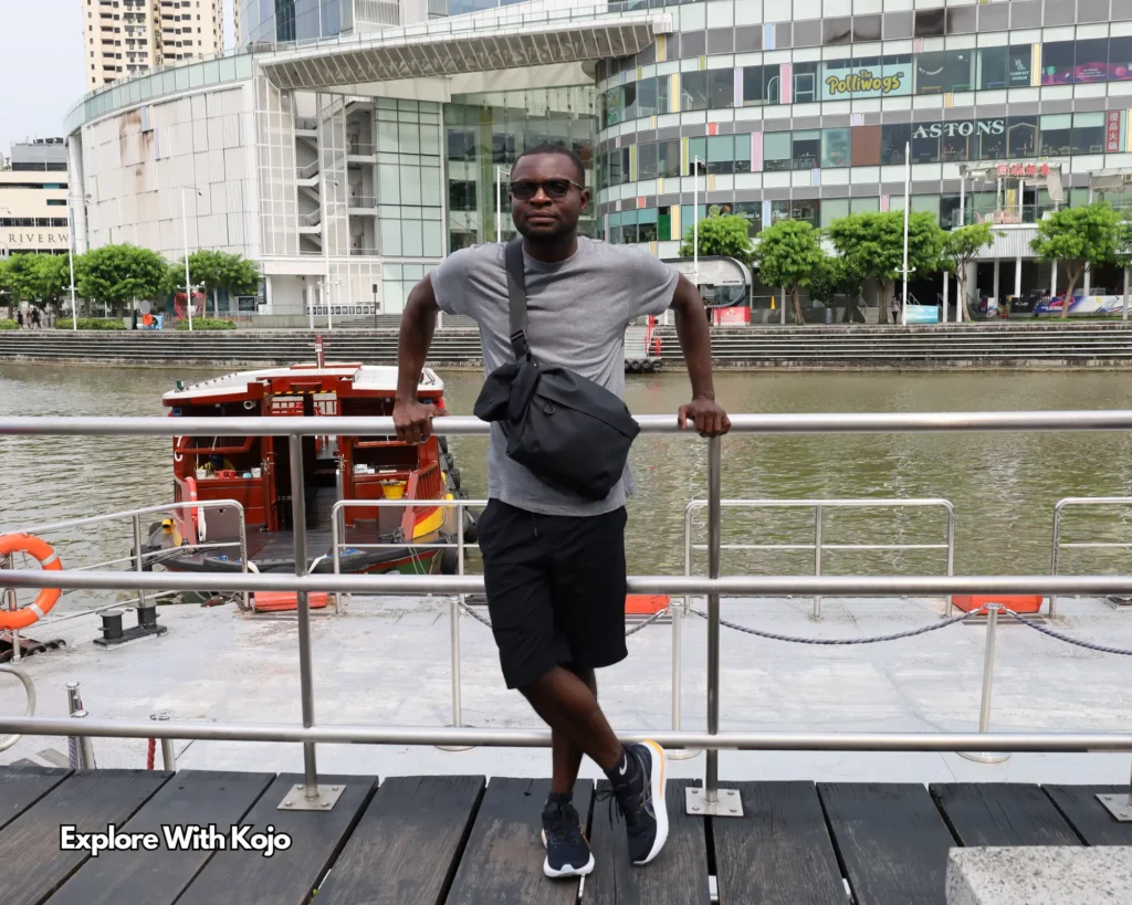 Kojo standing by the Singapore River with a traditional boat behind him, representing his travel experience in Singapore
