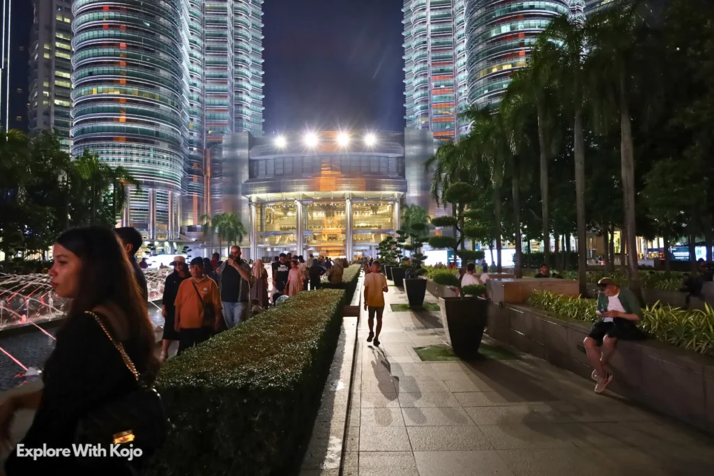 Night crowds along the KLCC Park walkway with the Petronas Twin Towers glowing in the background.