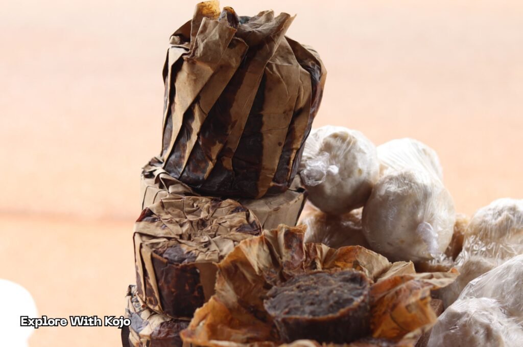 Stack of African black soap wrapped in cement-bag paper beside white ‘Tolon Banku’ soap balls wrapped in plastic, Ghana.