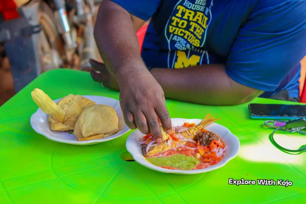 A man eating kenkey with fried fish, red pepper, and green sauce at a street food table.