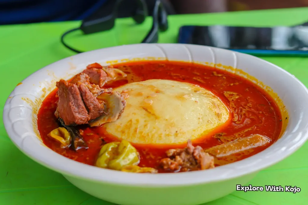 A bowl of palm nut soup with a mound of fufu, beef pieces, green chilies, and mushrooms.