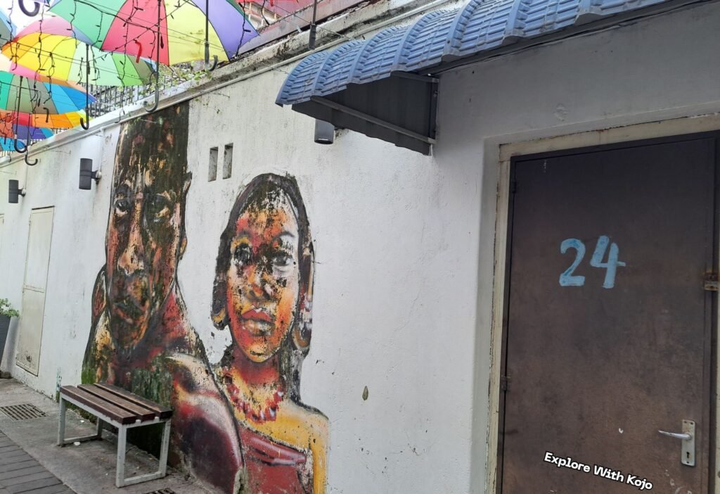 Photo taken by Explore With Kojo of street art in George Town, Penang, featuring a mural of two faces under colorful umbrellas.