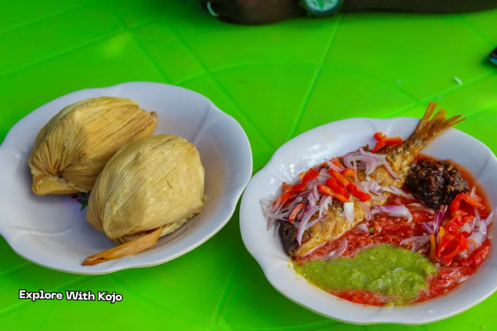 Two balls of kenkey served with fried fish, red pepper sauce, green chili, and onions.