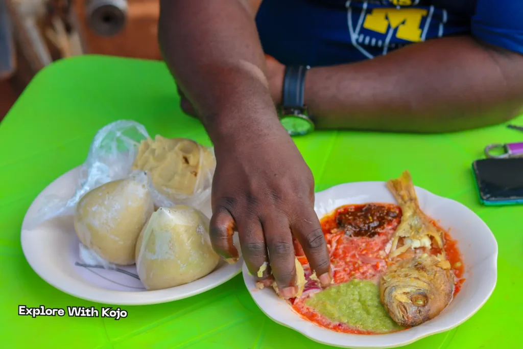 A man eating banku and fried fish with his hands, dipping into pepper sauce.