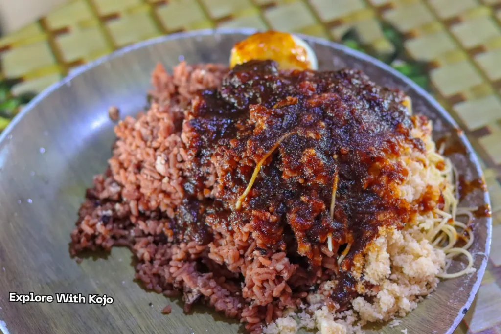 A Ghanaian waakye meal with boiled egg, fried fish, shito, and salad served in a plastic pack.