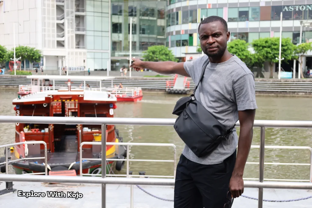 Kojo standing beside the Singapore River, pointing toward the river cruise boats at Clarke Quay.