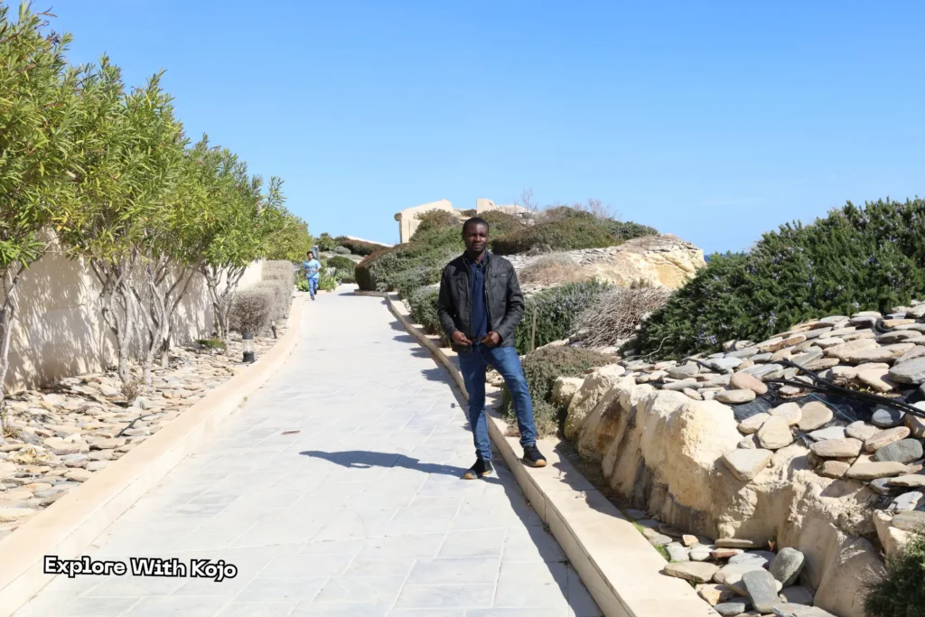 Kojo standing on a sunny coastal path in Malta, wearing a black jacket and jeans under a clear blue sky