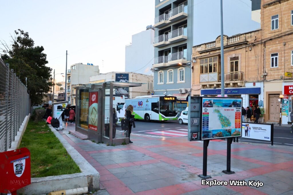 City bus stop in Sliema, Malta with bilingual signs, people waiting for the bus, and traditional Maltese buildings in the background