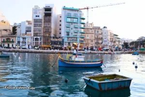 Colorful boats floating on the water in Malta with waterfront buildings and cafes in the background at sunset