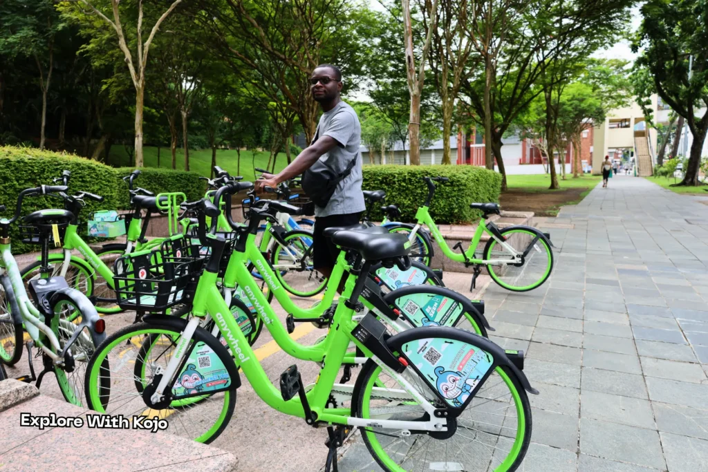 Kojo standing beside a row of bright green rental bicycles at Fort Canning Park, ready to explore Singapore.
