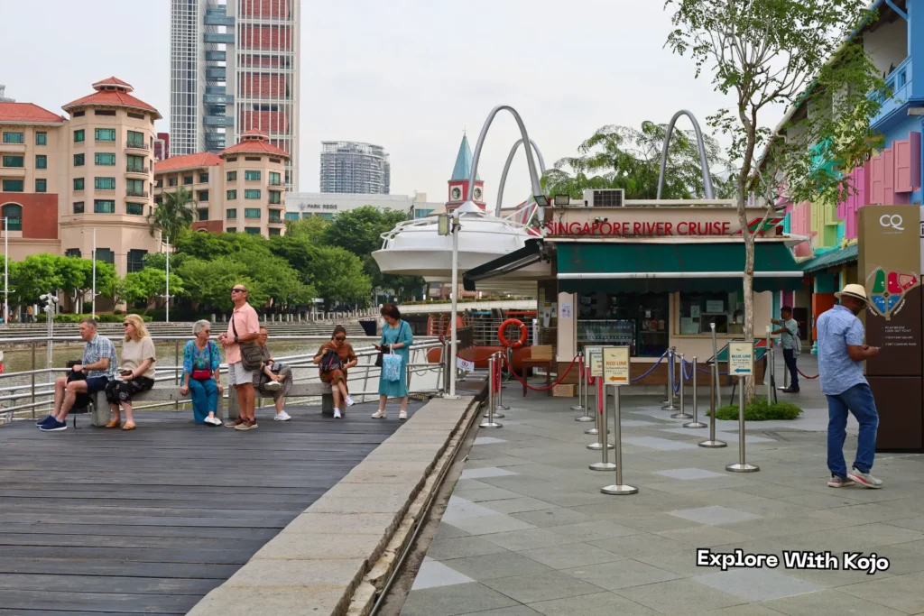 Tourists waiting at the Singapore River Cruise boarding point at Clarke Quay, with colorful buildings and boats along the riverside.