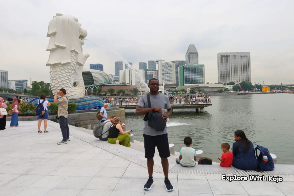 Kojo standing near the Merlion statue in Singapore with Marina Bay’s skyline in the background.