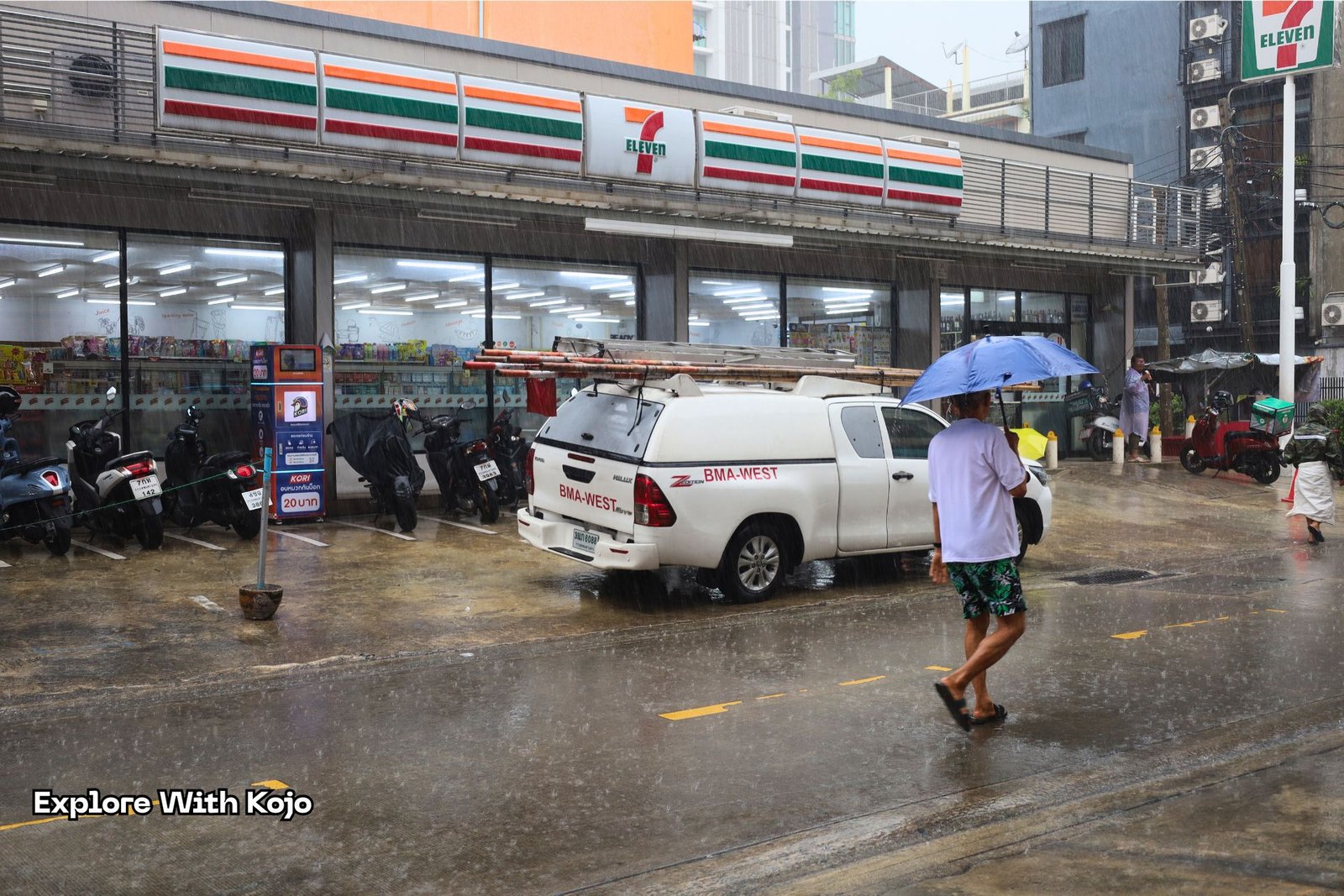 A Bangkok rainy day scene outside a 7 Eleven, showing a man walking with an umbrella during monsoon weather.
