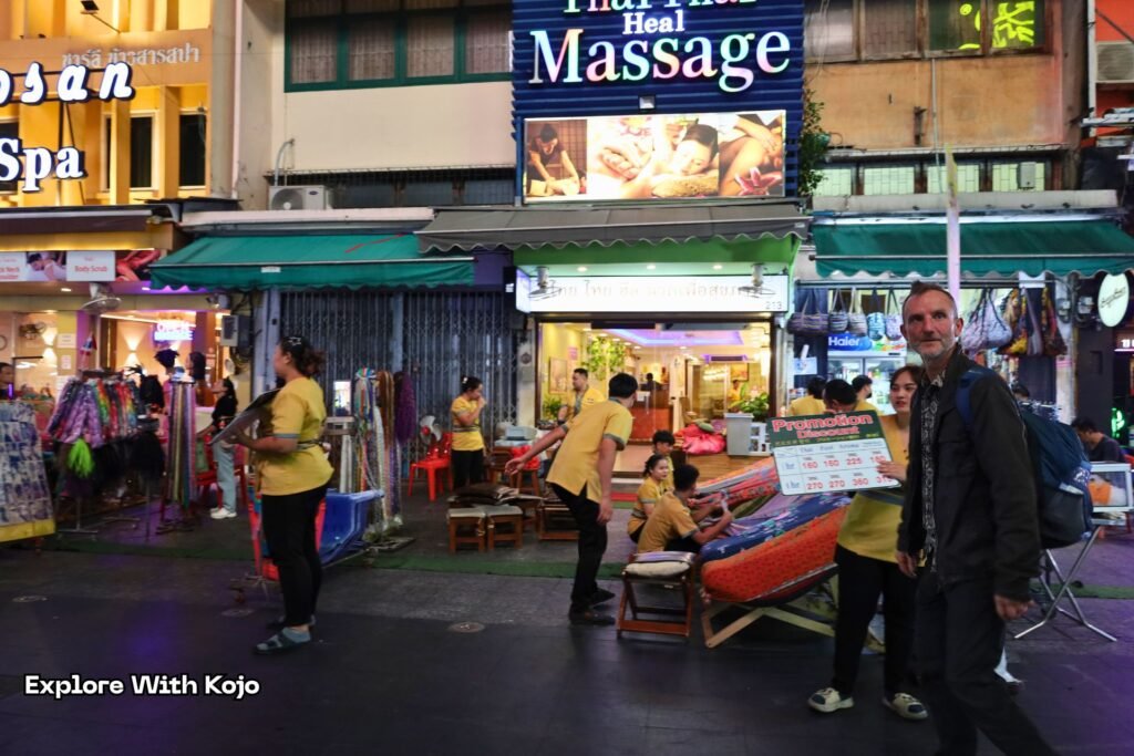 Street massage shops in Bangkok at night with workers offering foot massages and a traveler walking past.