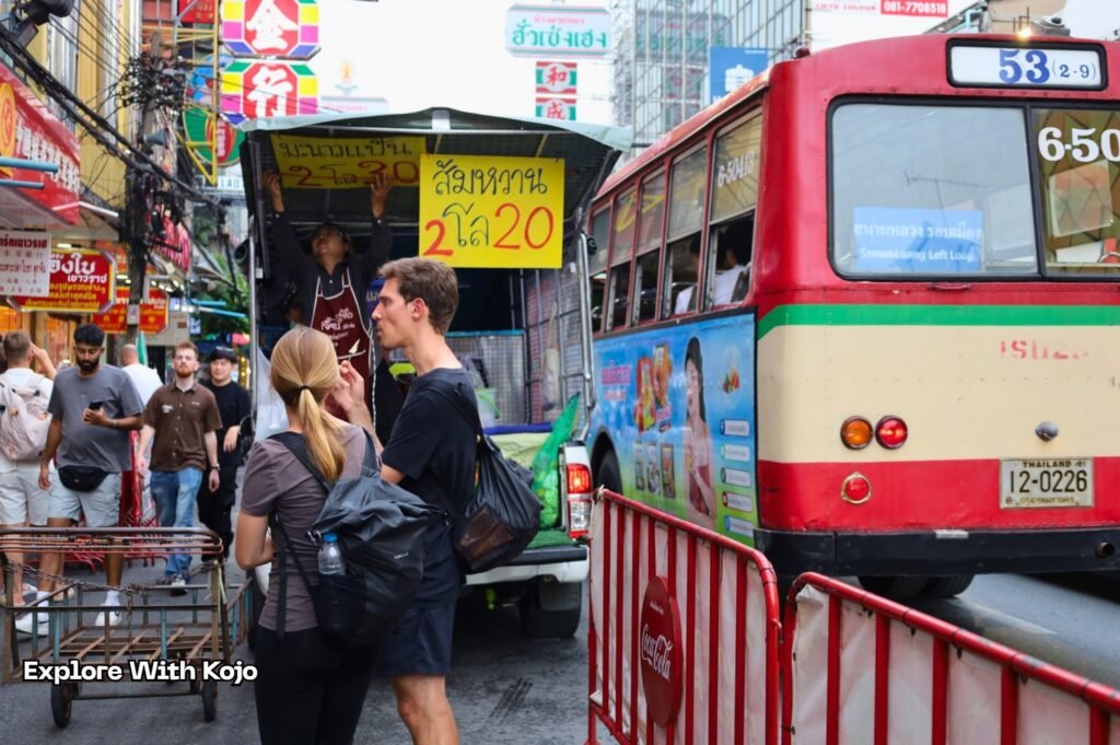Tourists walking through a busy street in Bangkok near a local bus and fruit stall, illustrating how Thailand’s new visa run crackdown affects everyday travelers.