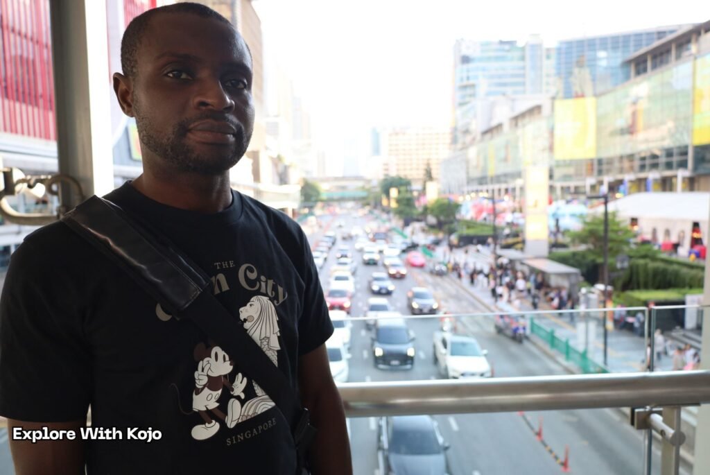 Kojo standing on a skywalk in central Bangkok with traffic below.