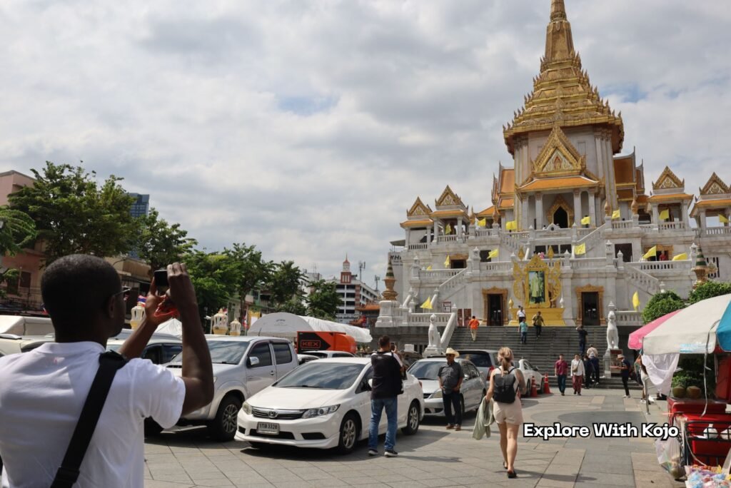 Kojo taking a photo outside Wat Traimit Temple in Bangkok, with the golden pagoda and visitors in the background.