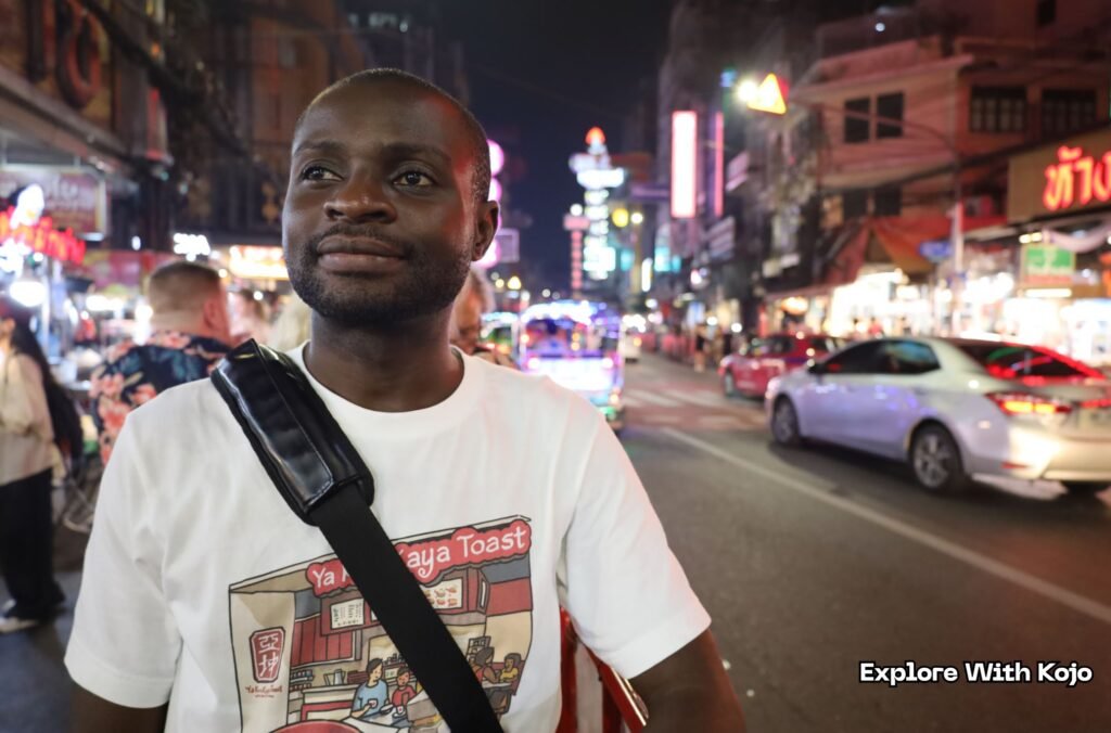 Kojo standing on a busy Bangkok street at night with neon lights and traffic in the background.