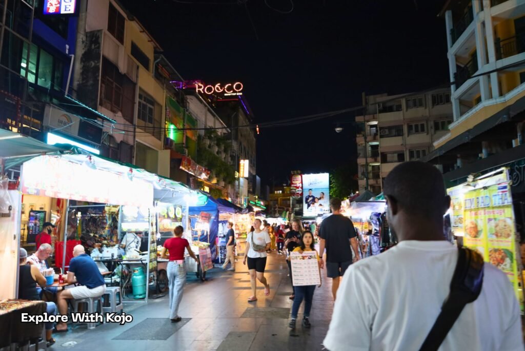 Kojo walking through Khao San Road at night, surrounded by street stalls, tourists, and bright lights in Bangkok.