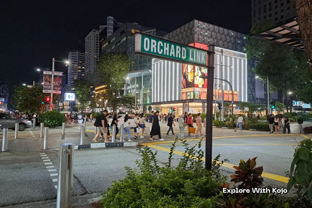 Night view of Orchard Road in Singapore showing the Orchard Link sign, luxury stores like Gucci, and people crossing the street under bright city lights.