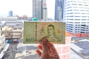 Hand holding a 20 baht Thai banknote with Bangkok city buildings in the background