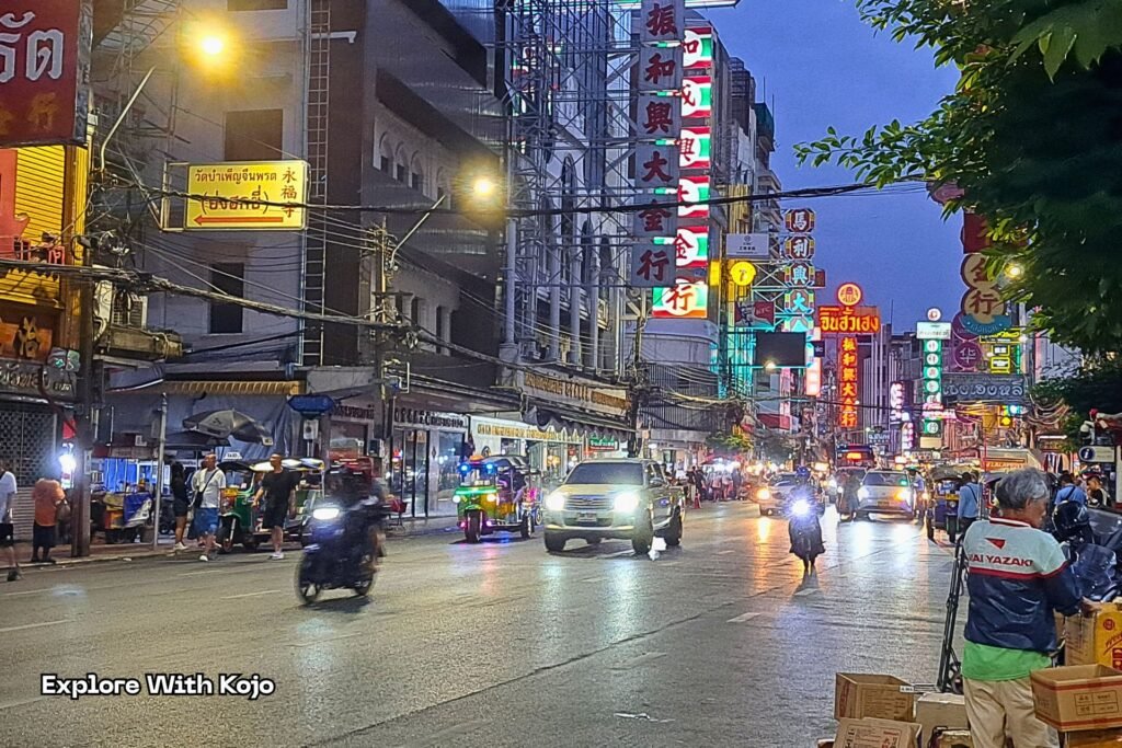 Night scene in Bangkok Chinatown with bright signs, traffic, and people on the street.